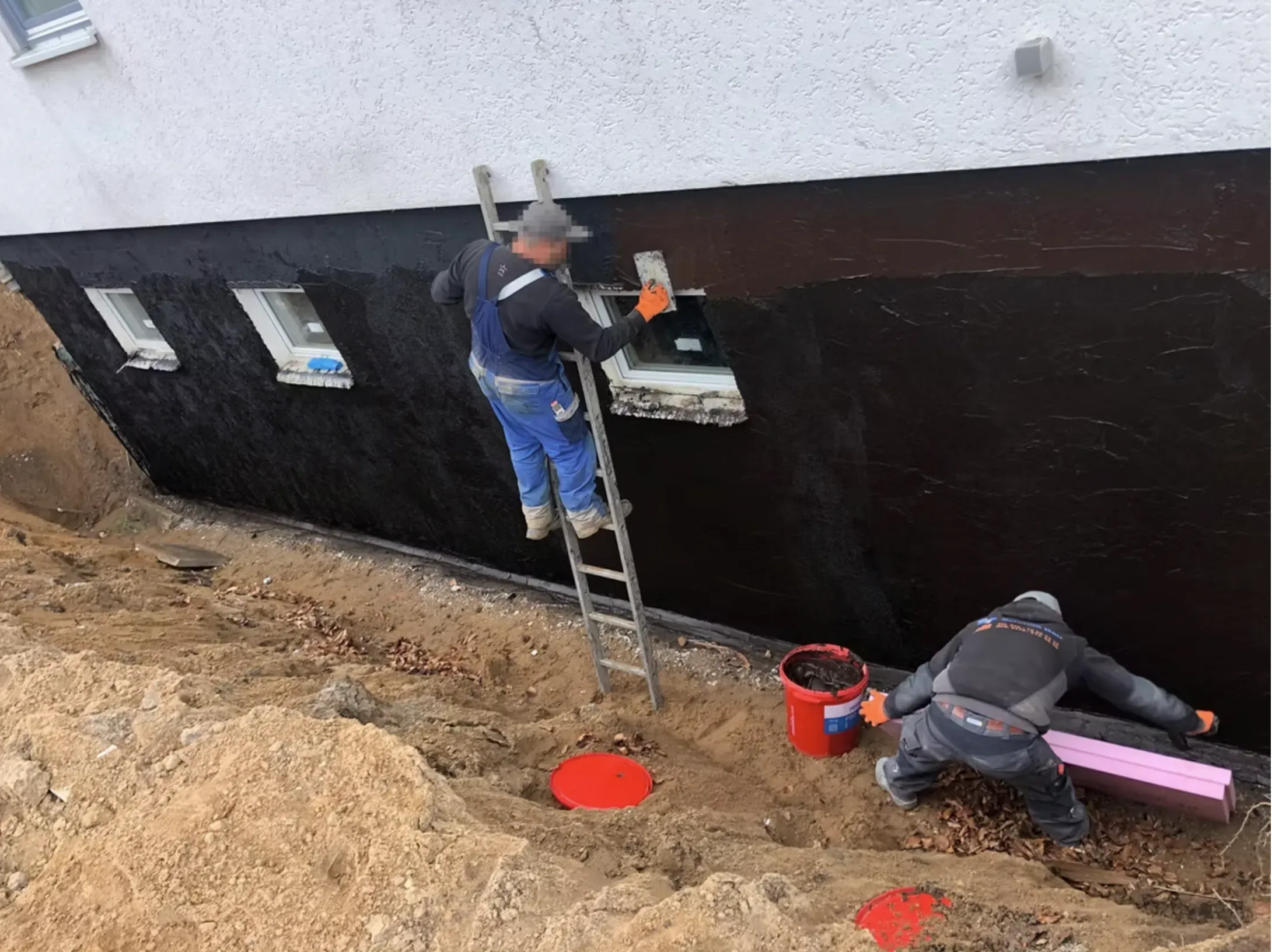 Freigelegte Kellerwand mit aufgetragener schwarzer Dickbeschichtung, davor zwei Kontrollschächte im Aushub und bereitgelegte Dämmplatten.