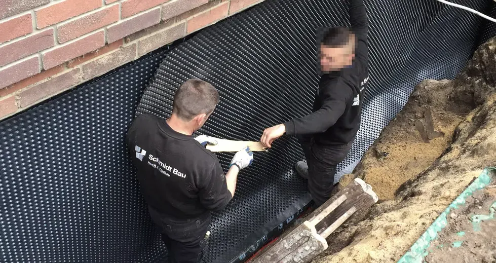 Zwei Mitarbeiter setzen in einer offenen Baugrube eine Noppenbahn auf die abgedichtete Kellerwand eines Ziegelbaus.