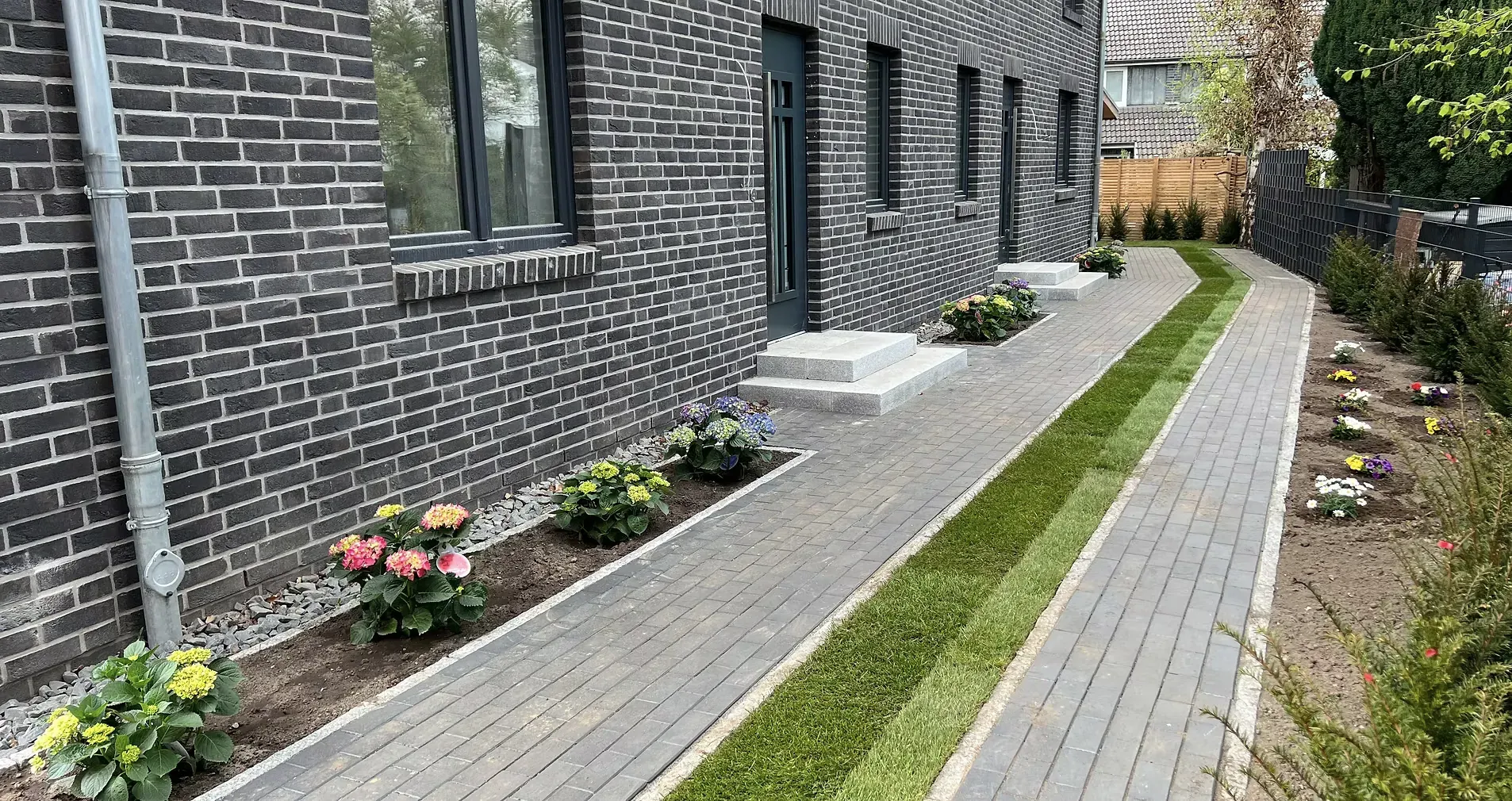 Neu gesetzter Pflasterweg entlang einer Klinkerfassade, daneben ein Pflanzstreifen und ein Rasenband.
